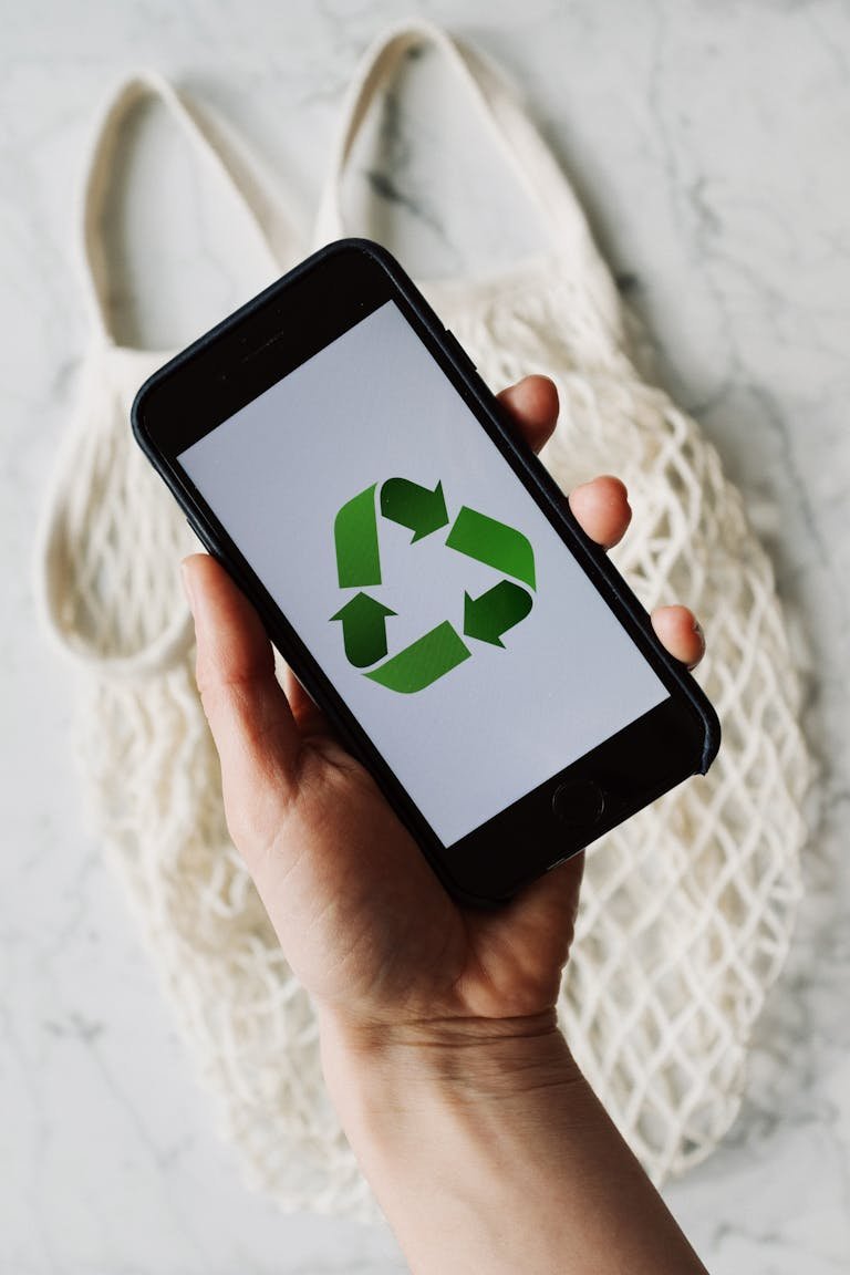 Top view of faceless person holding mobile phone with green recycle logo on white background above cotton eco mesh bag on marble table