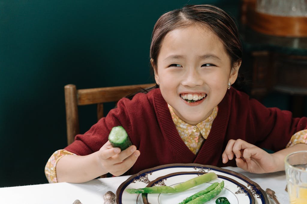 A happy child in a maroon sweater enjoying a meal with fresh vegetables at a dining table.