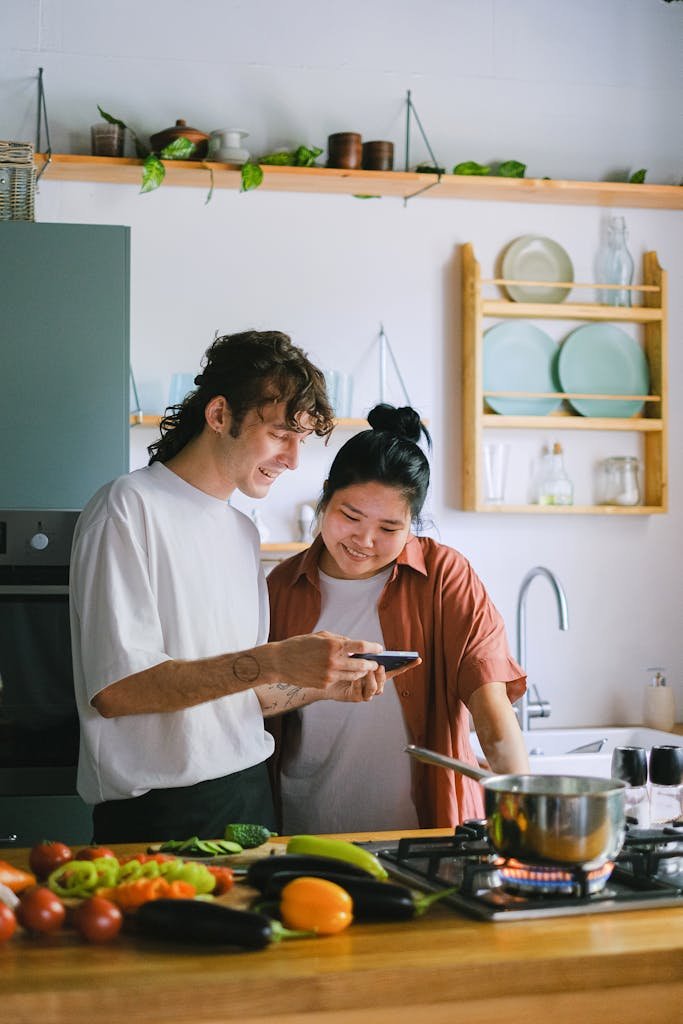 Happy couple using smartphone and preparing vegetables in modern kitchen. A moment of shared culinary joy.