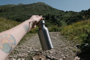A tattooed arm holding a stainless steel bottle against a scenic mountain backdrop.