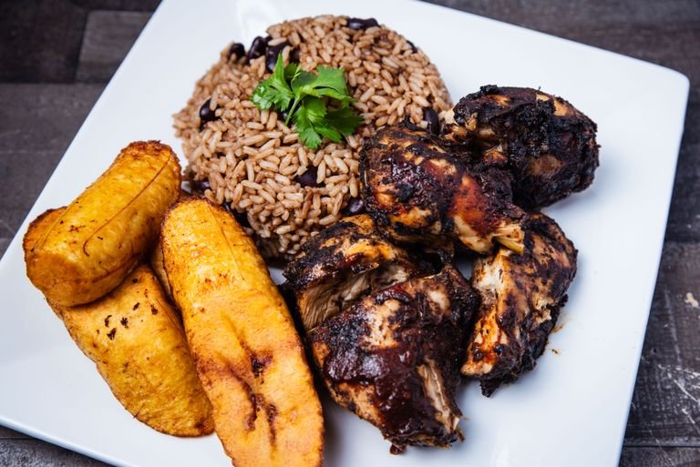 Tantalizing Caribbean jerk chicken served with rice and beans and fried plantains on a square plate.