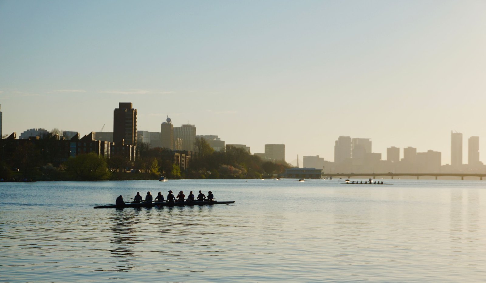 A team of rowers silhouetted on a calm river with a city skyline in the background during sunset.