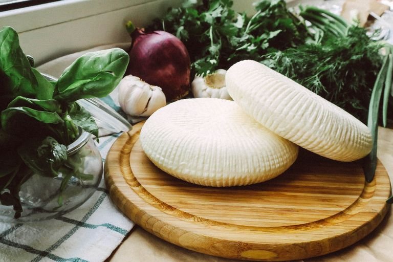 Delicious fresh ricotta cheese resting on a wooden board surrounded by basil, onion, and garlic.
