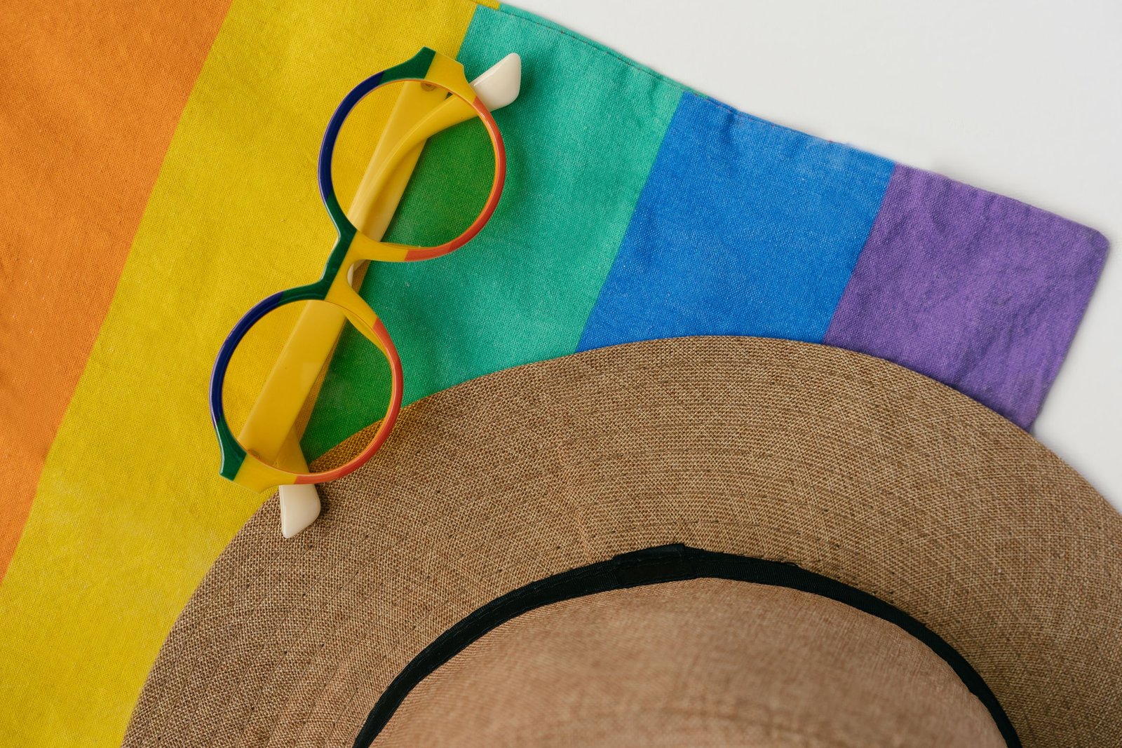 Flat lay of sunglasses and hat on a vibrant rainbow flag, capturing LGBTQ+ pride.