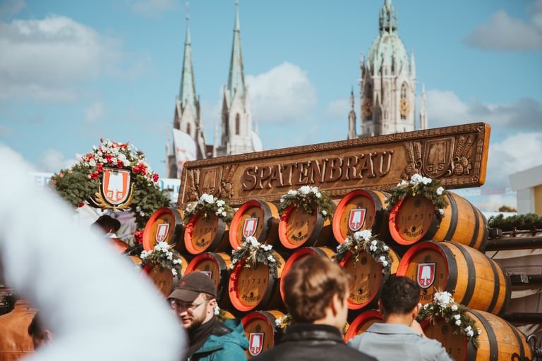 Vegan Oktoberfest in Munich scene at Spatenbräu, featuring beer barrels and festival-goers under a clear sky.